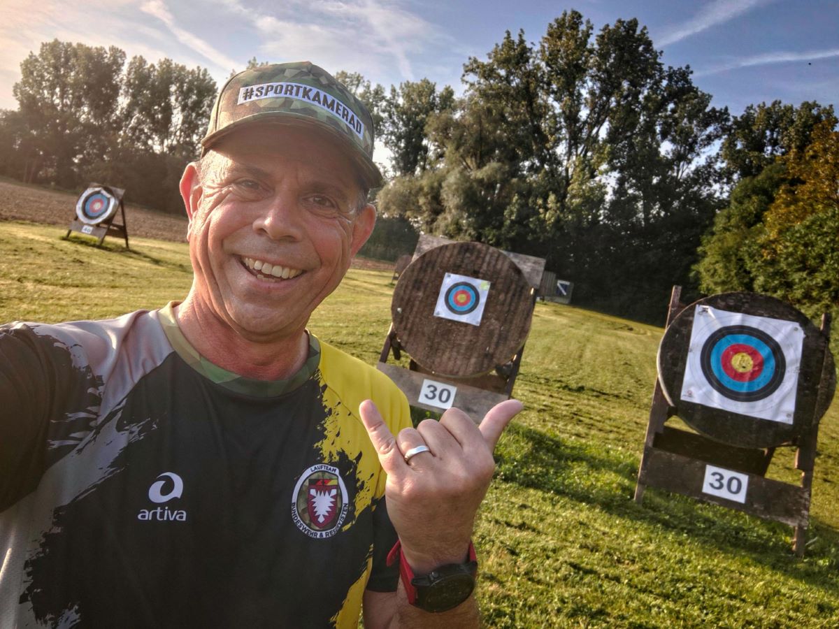 Sportkamerad Frank mit Shaka-Handzeichen auf einer Wiese, hinter ihm Zielscheiben für Bogenschießen